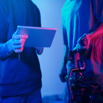 Dos personas colaborando en un entorno de producción multimedia con iluminación azul, una sosteniendo una tableta y la otra ajustando una cámara profesional